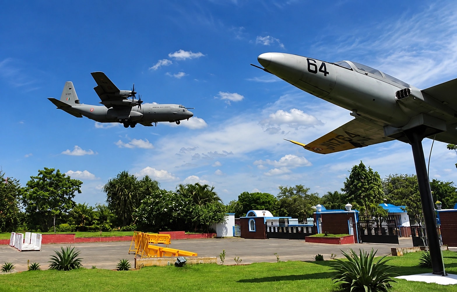 A preserved jet mounted on a plinth beside the gates of an air force station, a transport aircraft coming in to land above it
