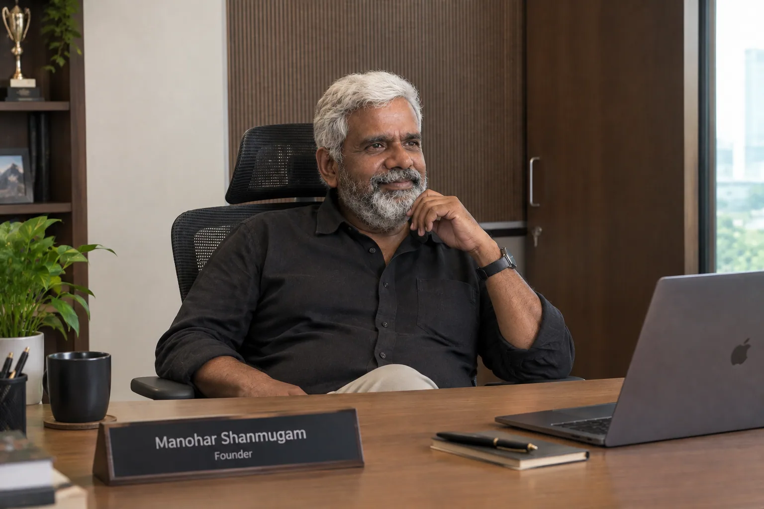 Manohar Shanmugam smiling at his office desk in a black shirt, laptop open before him and a city view through the window behind
