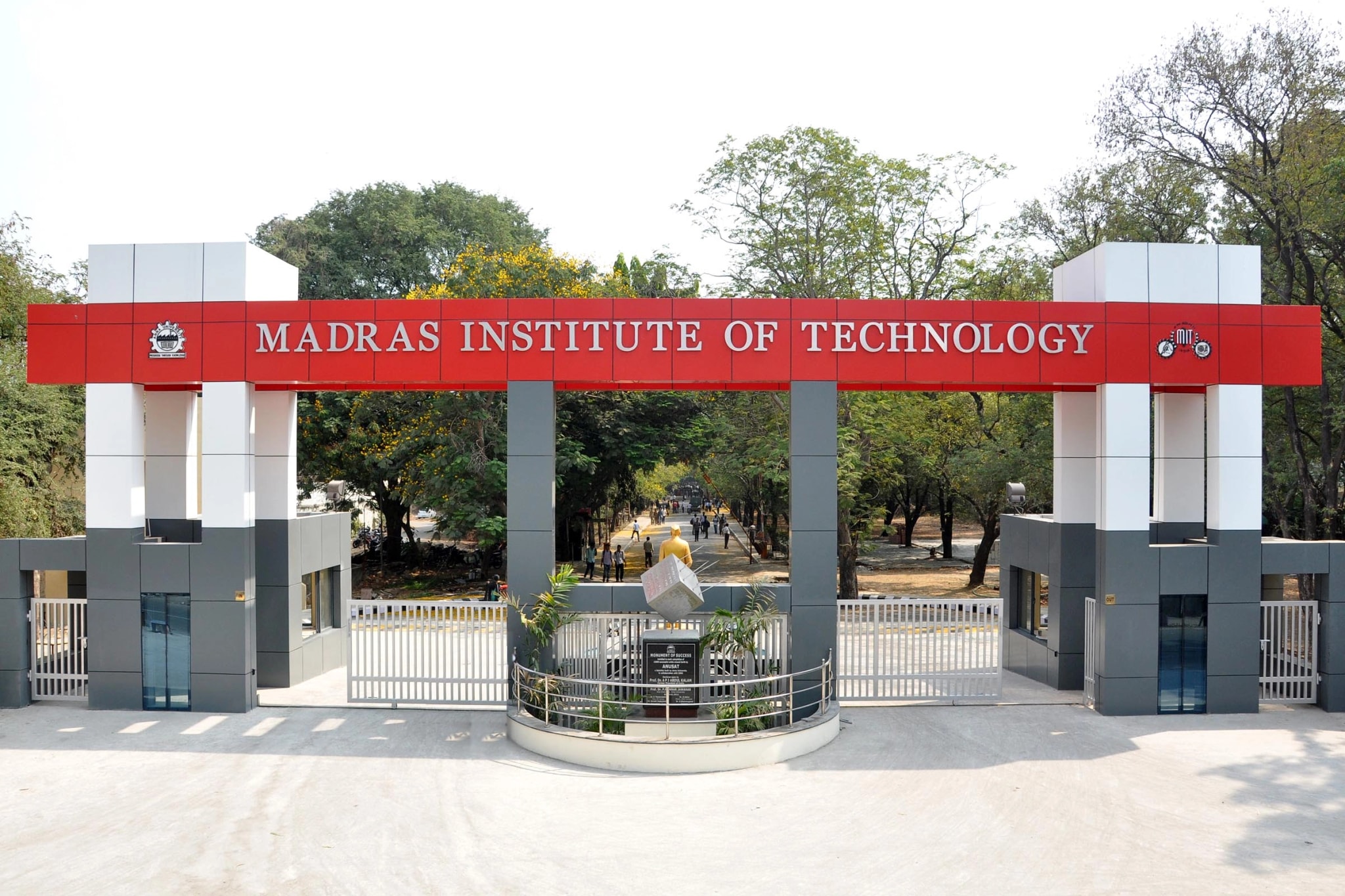The Madras Institute of Technology entrance gate, its red sign spanning white and grey pillars, with a tree-lined avenue beyond