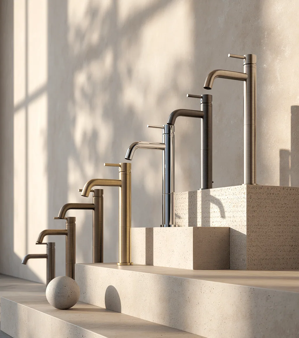 Basin faucets in brushed gold, chrome and gunmetal finishes displayed on plinths against a sunlit stone-textured wall