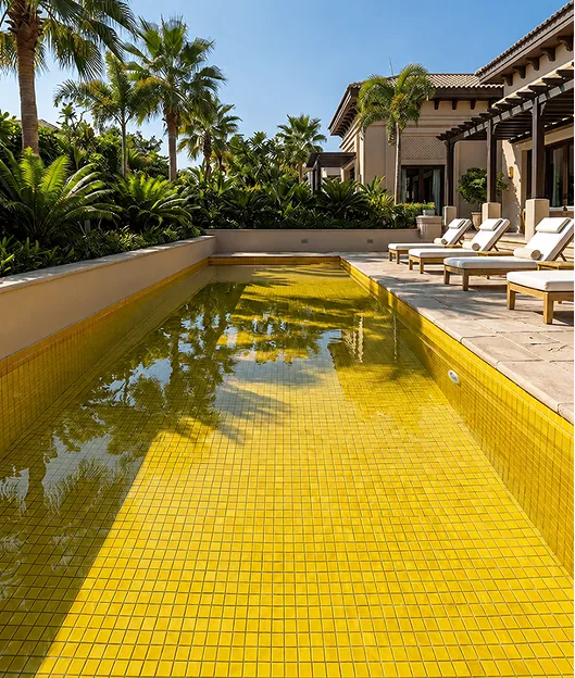 Swimming pool lined in yellow mosaic tile, with beige stone paving and palm trees around it